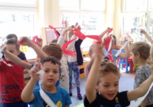 Grupa dzieci podczas zabawy ruchowej, wymachują biało-czerwonymi paskami bibuły.